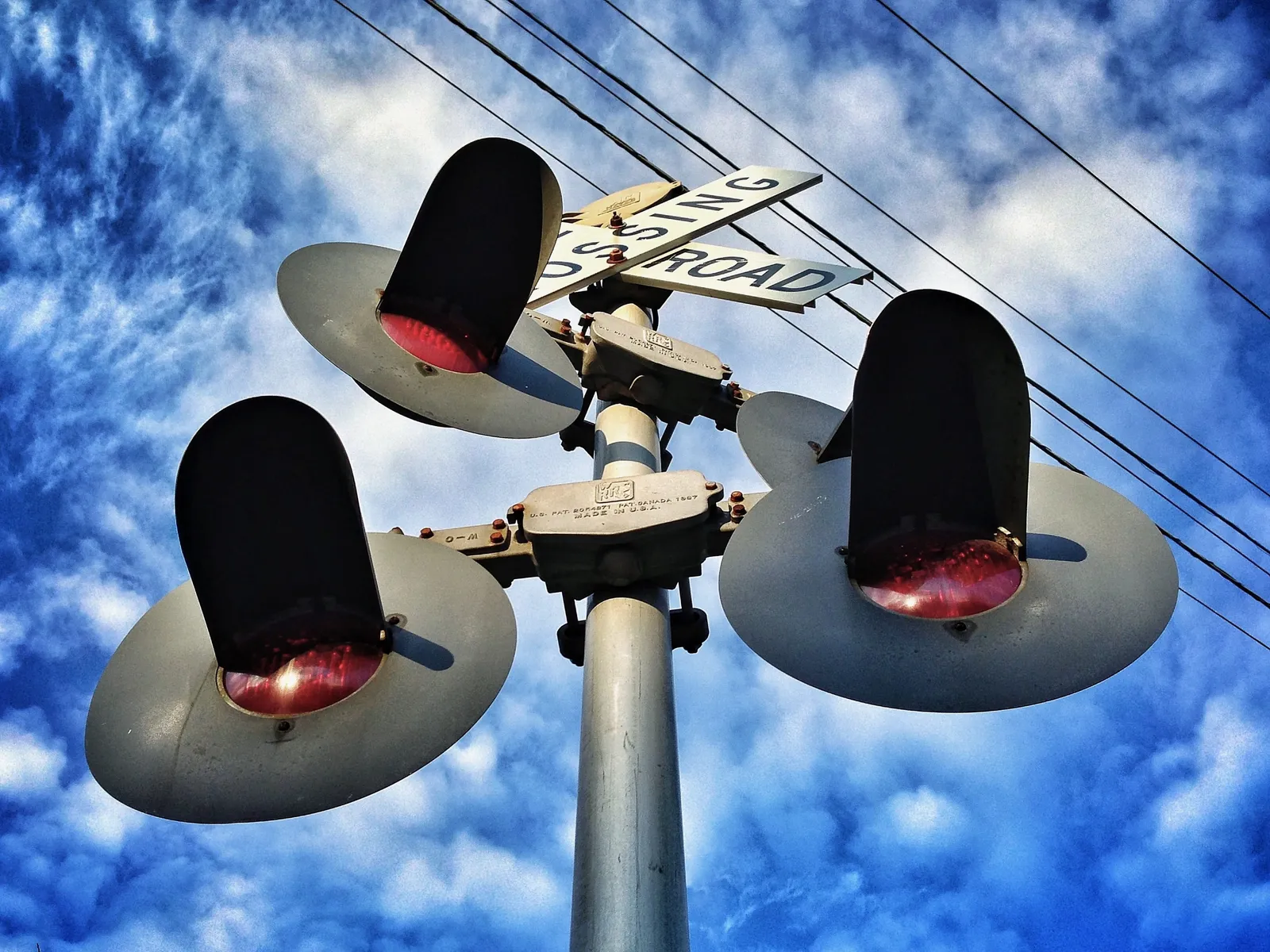 Looking up at a railroad crossing sign against a blue sky with some wispy clouds. Some power lines cut diagonally across the background between the sign and the sky.