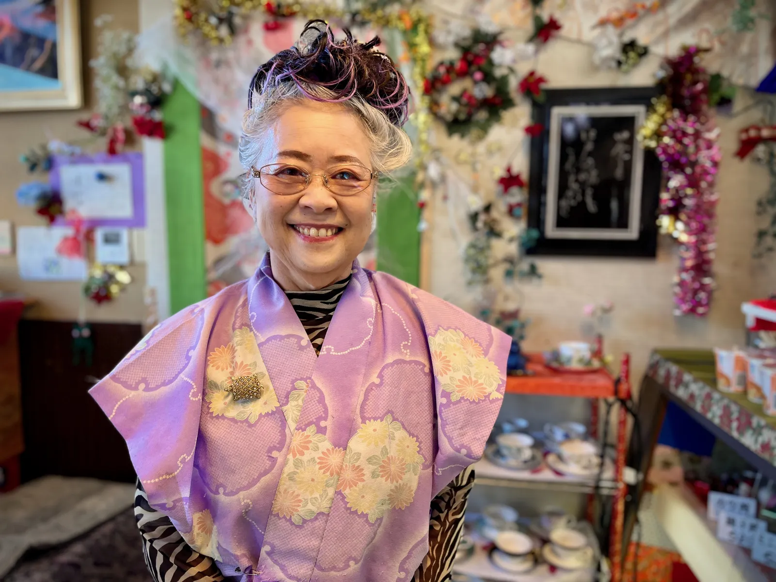 An older woman standing in the dining room of a small sushi restaurant smiles broadly for the camera.
