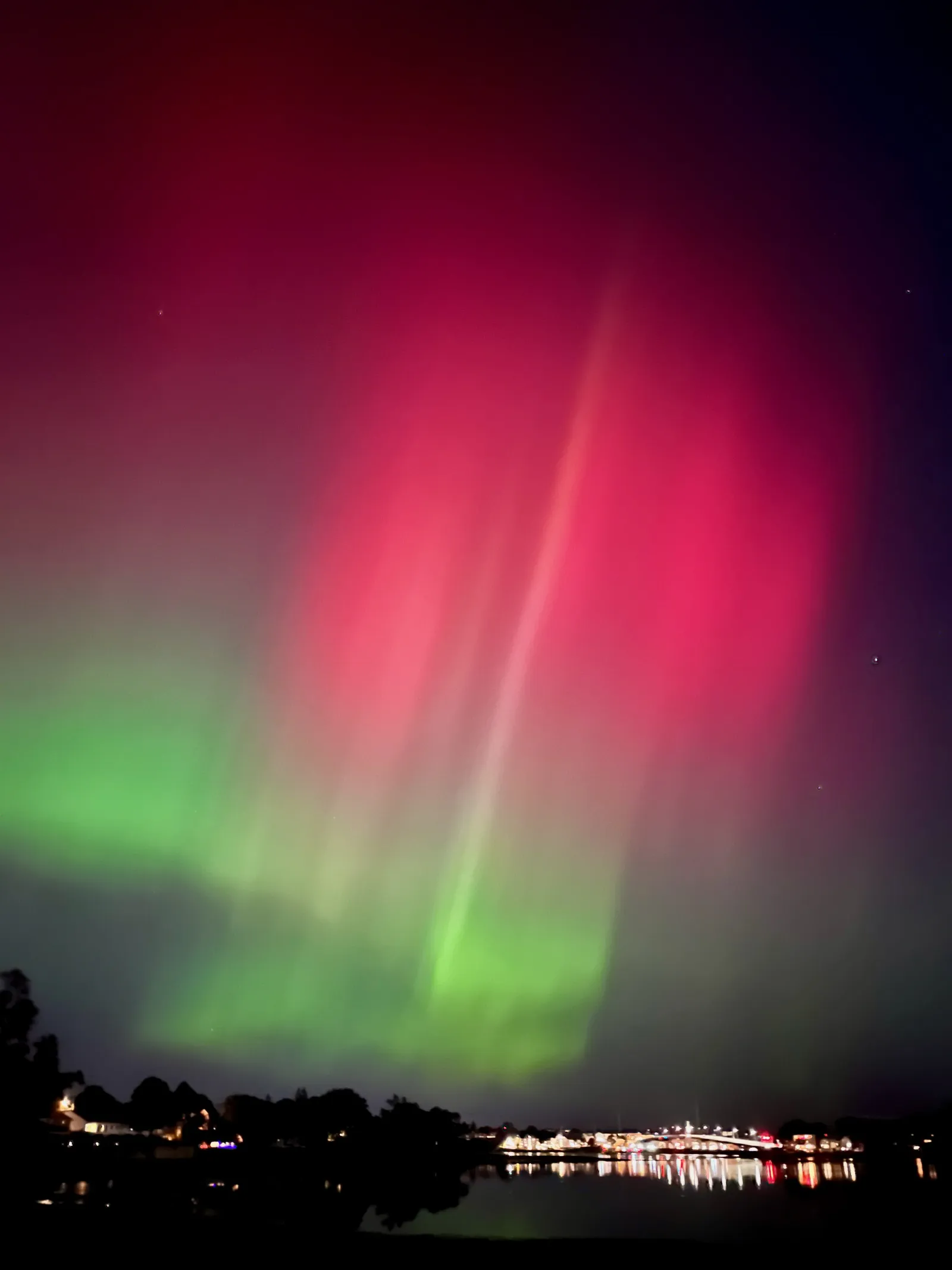 Green and red aurora light up the sky over a body of water. In the background, along the horizon, we can see some city lights and a bridge.